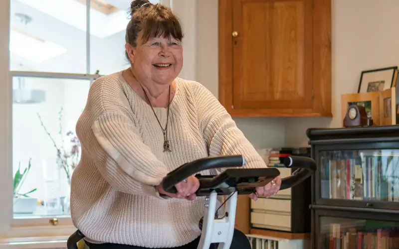 Lachende oudere vrouw fietst op een witte opvouwbare hometrainer in haar woonkamer